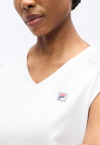 Λευκό τοπ με V λαιμόκοψη και κοντά μανίκια. Στην αριστερή πλευρά του στήθους υπάρχει ένα τετράγωνο λογότυπο με κόκκινες και μπλε λεπτομέρειες. Λεία υφή με άνετη εφαρμογή.