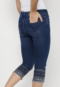 Pantalons capri en denim bleu foncé avec des coutures décoratives et des poignets brodés aux accents motifs et un ourlet effiloché.