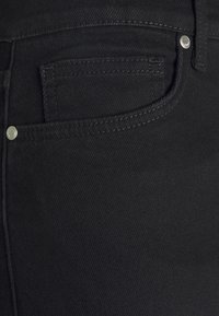 Gros plan d'une poche de jean en denim noir avec des coutures visibles et deux rivets métalliques sur le bord supérieur.