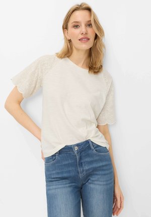 Lyshåret kvinde iført en hvid kortærmet top med øjeskårede blondeærmer og blå højtaljede jeans, poserer med den ene hånd på hoften.