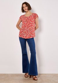 Blusa roja de flores con mangas cortas, escote redondo y detalles fruncidos. Combinada con jeans de campana azules y sandalias de tacón cuadrado marrones.