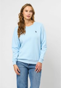 Ljusblå sweatshirt med lång ärm, rund halsringning och en liten svart logotyp på bröstet. Texturerat tyg, avslappnad passform. Kombinerad med denimjeans.