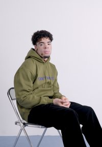 Sweatshirt com capuz verde oliva e logótipo roxo, calças pretas, sentado numa cadeira prateada contra um fundo branco simples.