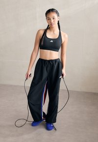 Svart sports-bh med logo, løse svarte joggebukser med røde, hvite og blå sidelabber, og blå joggesko som holder et hoppetau.