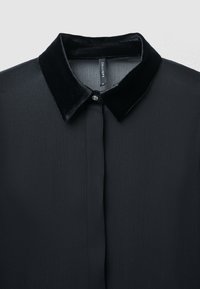 Camicia scura in tessuto morbido e testurizzato, con colletto in velluto nero e un dettaglio a bottone unico sul colletto.
