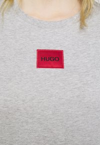 Grå bomulds t-shirt med en rød rektangulær lap med teksten "HUGO" i sort. Stoffet har en glat tekstur med en klassisk halsudskæring.