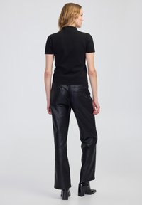 Kobieta z jasnymi włosami stoi, ubrana w czarny top z krótkim rękawem, czarne skórzane spodnie i czarne botki na obcasie, odwrócona tyłem na jednolitym tle.