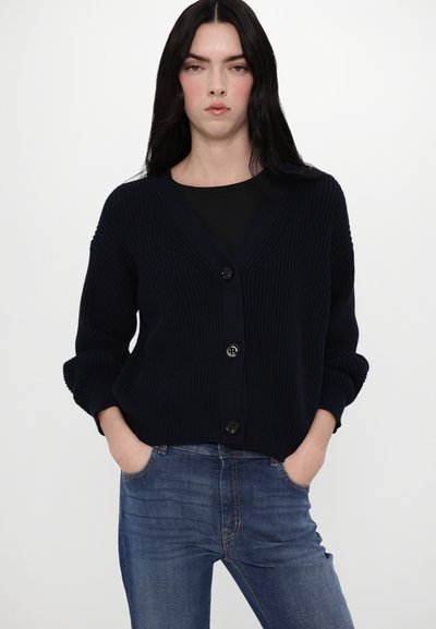 Navy-blauer gerippter Cardigan mit V-Ausschnitt, vier großen Knöpfen, überschnittenen Schultern und hochgekrempelten Ärmelbündchen, kombiniert mit blauen Jeans.