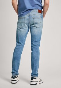Vaqueros de denim azul claro con un corte entallado, diseño de cinco bolsillos y parche de piel en la parte trasera de la cintura. Usados con zapatillas blancas y marinas.