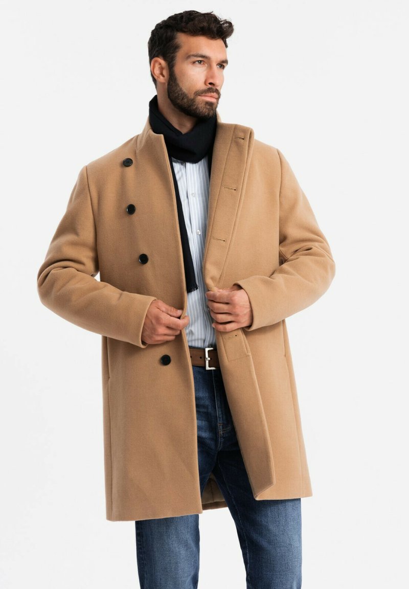 Kamelfarbener Mantel mit einem Doppelreiher-Design und schwarzen Knöpfen. Getragen über einem hellblau gestreiften Hemd, kombiniert mit blauen Jeans und einem schwarzen Schal.