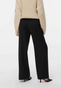 Pantalons larges noirs en tissu doux associés à un gros pull en tricot beige. La tenue présente des détails de manches côtelées et une coupe décontractée.