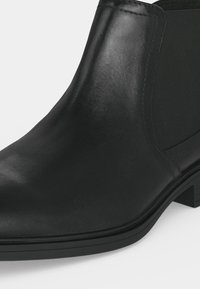 Bottine en cuir noir avec bout arrondi, panneau latéral élastique et petit talon empilé sur fond blanc.