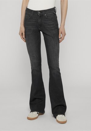 Personne portant un jean bootcut noir et des baskets blanches, debout devant un fond gris clair uni.