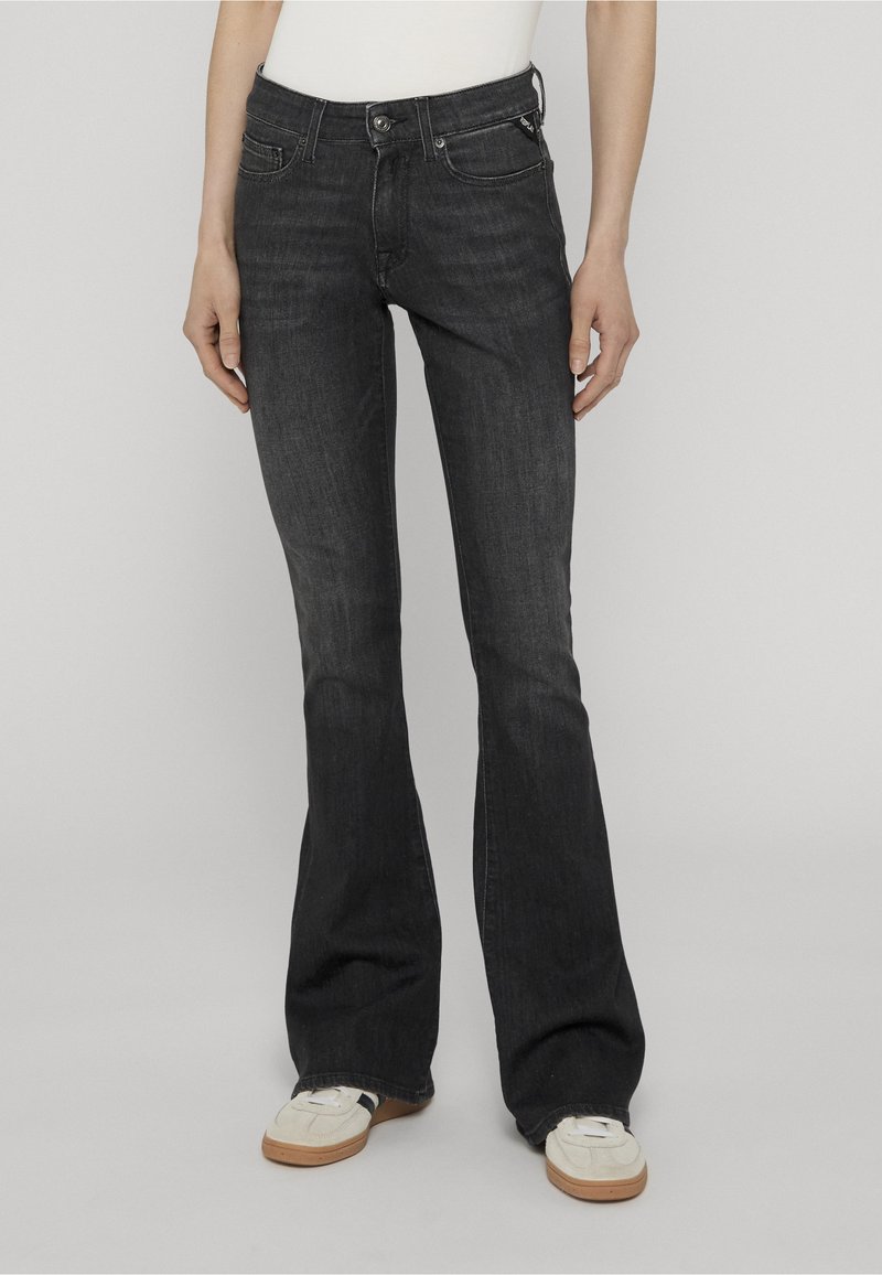 Άτομο φοράει μαύρα τζιν bootcut και λευκά αθλητικά παπούτσια, στέκεται μπροστά σε απλό ανοιχτογκρι φόντο.