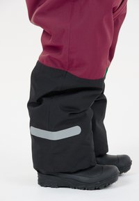 Waterdichte broek in bordeauxrood en zwart. Voorzien van een grijze reflecterende strip. Gemaakt van flexibele, gestructureerde stof. Ontworpen voor gebruik buitenshuis.