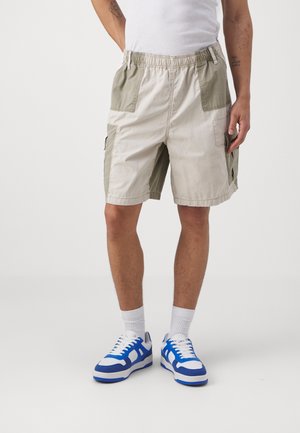 Homme portant un short cargo beige et vert olive clair, un débardeur blanc, des chaussettes blanches et des baskets bleues et blanches, debout devant un fond uni.