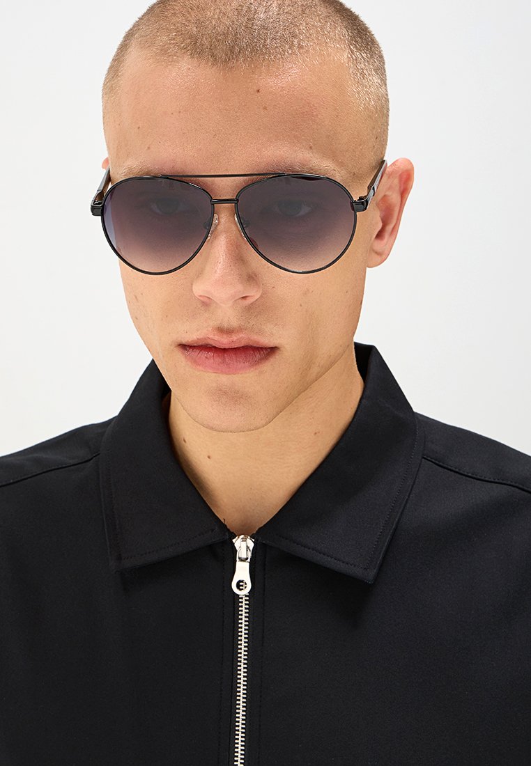 Occhiali da sole rotondi con lenti a gradiente, montatura in metallo nero, indossati con una giacca nera con zip. Texture liscia, dettagli minimi.