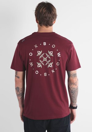 T-shirt en coton bordeaux avec un graphisme circulaire orné de motifs floraux et le mot "OXBOW" en blanc, à manches courtes et col rond.