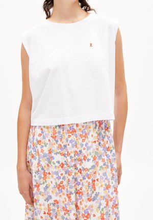 Frau trägt ein weißes, ärmelloses Crop-Top mit einem kleinen orangen Logo und einen hoch taillierten Rock mit roten, lila, gelben und grünen Blumenmustern.