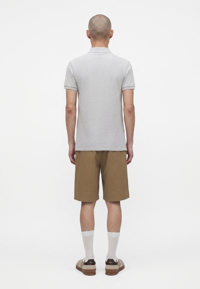 Homme debout de dos portant un polo gris clair à manches courtes, un short marron, des chaussettes blanches et des chaussures beiges sur fond blanc.