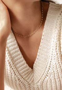 Strickpullover in Creme mit tiefem V-Ausschnitt, versehen mit strukturierten Maschen, kombiniert mit einer zarten goldenen Kettenhalskette mit ovalen Gliedern.