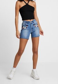 Shorts in denim azzurro chiaro con motivi floreali ricamati sulle tasche anteriori. Abbinati a un top nero corto e sneakers bianche.