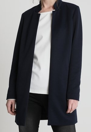 Person iført en lang, åben navy blazer over en hvid top og sorte bukser, stående mod en ensfarvet lysegrå baggrund.