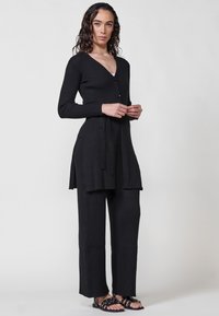 Conjunto de punto negro con canalé, escote en pico, dos botones, aberturas laterales y cinturón en la cintura. Combinado con sandalias de dedo al descubierto con acentos de metal.