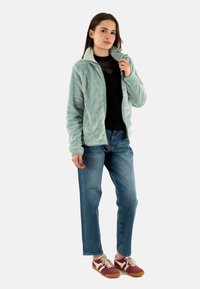 Chaqueta de forro polar texturizada en verde claro con cuello alto, llevada sobre una camisa negra, combinada con jeans azules y zapatillas rojas-borgoña.
