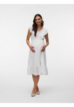 Tehotná žena s dlhými tmavými vlasmi, oblečená v biele midi šaty bez rukávov s dierkovaným vzorom a béžovými balerínkami, usmievajúca sa, stojaca s rukami na bruchu.