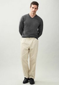 Grau v-neck Pullover, hellbeige plisierte Hose, schwarze Schuhe. Strickstruktur auf dem Pullover; die Hose ist glatt und locker geschnitten.
