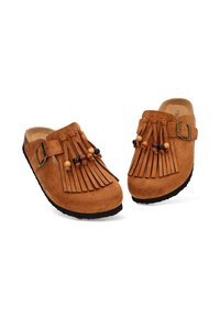 Zuecos de ante en marrón claro con detalle de flecos en la parte delantera, adornados con cuentas de madera. Suelas de goma negra con hebilla ajustable.