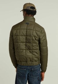 Olivgrön puffjacka med ett kviltat mönster, hög krage och elastiska ärmslut. Har en liten logopatch vid nacken.