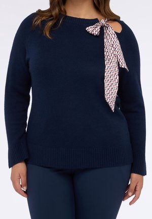 Kobieta nosząca granatowy sweter z wycięciem na ramieniu i ozdobną kokardą z materiału o wzorze, zawiązaną przy dekolcie.