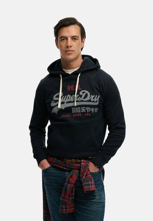 Młody mężczyzna w czarnej bluzie z kapturem SuperDry z białymi sznureczkami i czerwoną kratowaną koszulą zawiązaną wokół talii.