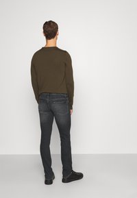 Hombre de espaldas con suéter verde oliva, jeans gris oscuro de corte slim y zapatillas negras contra un fondo claro y liso.