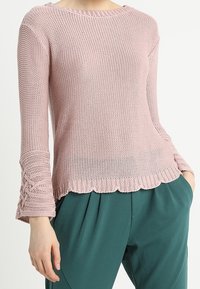 Ljusrosa, stickad tröja med lång ärm, vågig nederkant och texturerade detaljer på ärmarna. Matchas med gröna byxor.