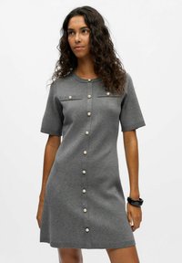 Robe en tricot grise à manches courtes, avec des détails de boutons en perles sur le devant et deux poches poitrine. Texture douce et design ajusté.