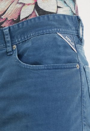 Niebieska kieszeń dżinsów z denimu z widoczną metką marki Replay oraz częścią wzorzystego kwiatowego topu nad linią talii.