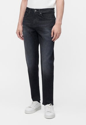 Personne portant un jean slim gris foncé et des baskets blanches, debout devant un fond clair uni.