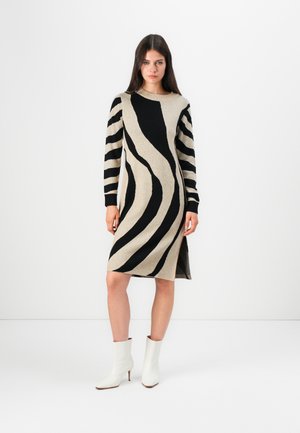 Robe en tricot avec une base beige et des motifs ondulés noirs, dotée de longues manches rayées et d'une fente sur le côté. Associée à des bottines blanches.
