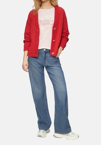 Rode gebreide cardigan met knoopsluiting, gedragen over een lichtroze shirt met tekst. Gecombineerd met blauwe brede denim jeans en witte sneakers.