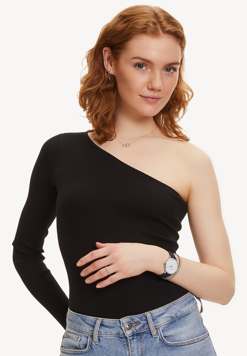 Zwart geribbeld bodysuit met één lange mouw en één blote schouder. Model draagt lichtblauwe jeans en een zilveren horloge, met golvend haar.