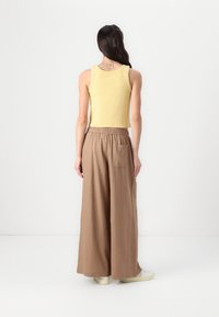 Débardeur jaune côtelé et pantalon large marron avec une taille élastique. Le mannequin porte des baskets blanches, avec un tatouage visible dans le dos.