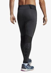 Men's black athletic leggings with textured mesh panels, and a zippered pocket at the waistband. Worn with blue athletic shoes.