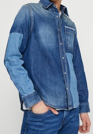 Mężczyzna w koszuli z dżinsu patchwork i niebieskich dżinsach, z jedną ręką w kieszeni, na jednolitym tle.