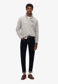 Sudadera gris con cuello de media cremallera y logo impreso, combinada con jeans oscuros y zapatos marrones, con los puños doblados y un reloj.