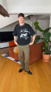 Junger Mann trägt ein schwarzes T-Shirt mit Grafik und olivgrüne Hose, steht im Wohnzimmer mit Holzboden, orangefarbenem Sofa, großer Pflanze und Fernseher im Hintergrund.