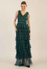 Langes, gestuftes Abendkleid aus tiefgrüner Tüll, mit V-Ausschnitt, Taillengürtel und geschichteten Rüschen, kombiniert mit schwarzen High Heels.