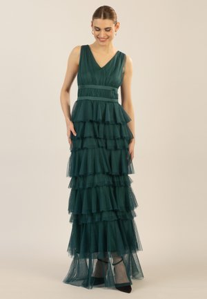 Long, tiered evening dress in deep green tulle, featuring a V-neckline, waist belt, and layered ruffles, paired with black heels.
